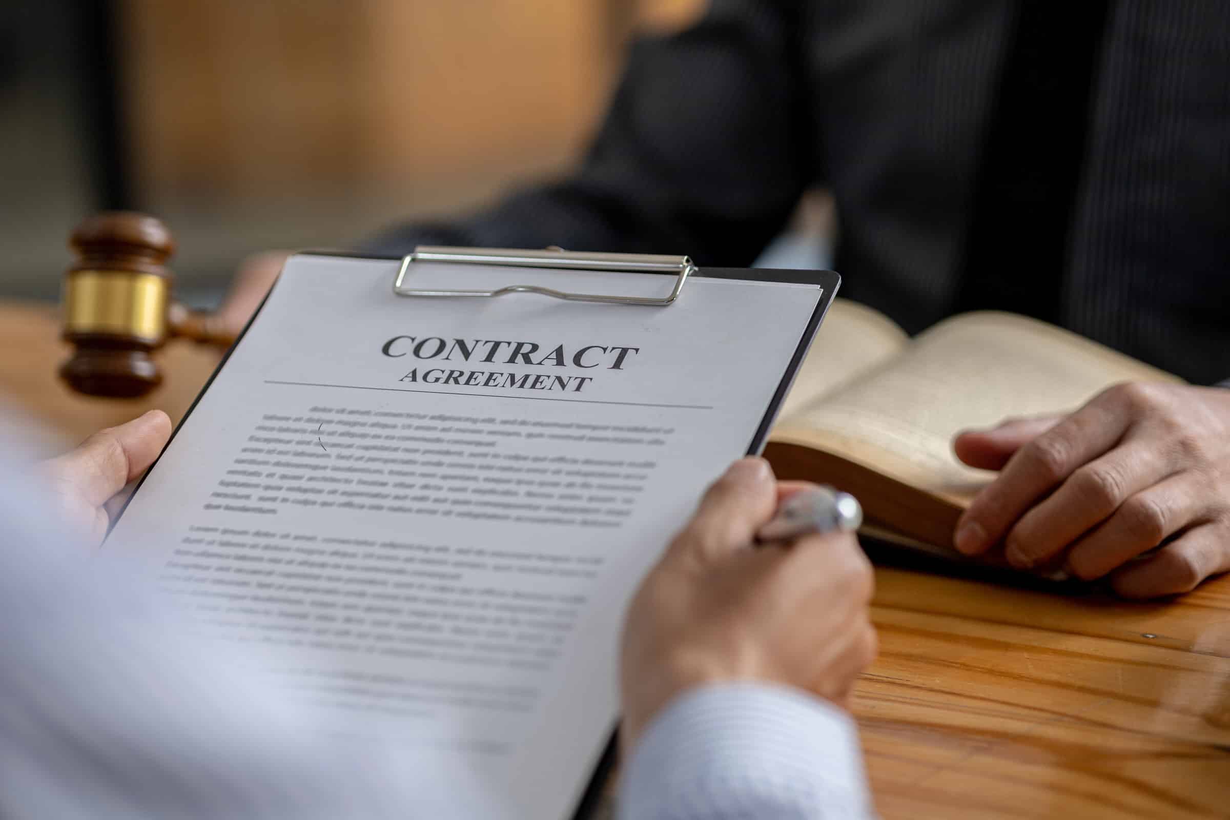 A person holds a clipboard with a breach of contract agreement while another sits across the table; a judges gavel hints at civil litigation nearby.