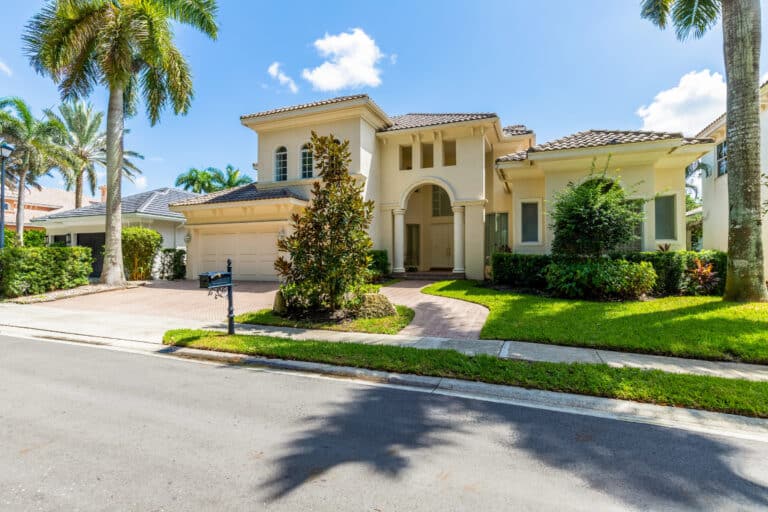Two-story white stucco house with a tile roof, arched entryway, and palm trees—an inviting example of Florida real estate viewed from the street on a sunny day.